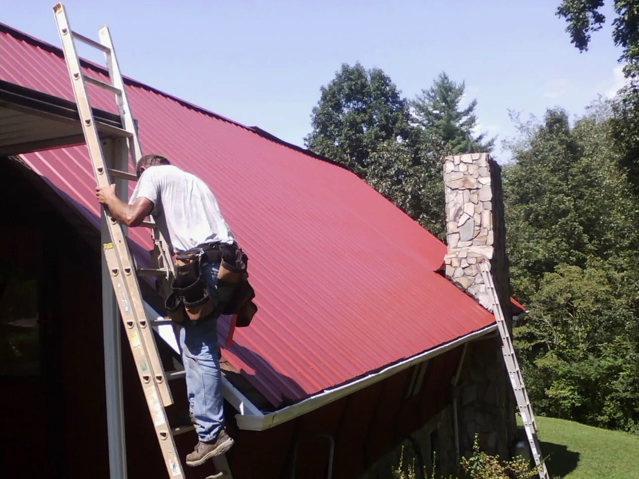 Roofer on a ladder beside a red metal roof and stone chimney — Soffit & Fascia Repair by Roofing Brothers in Hayden